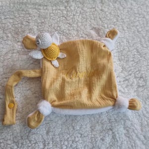 Peut inclure: Une couverture de confort jaune pour bébé avec une tortue crochetée attachée. La couverture a une bordure blanche et le nom "Carmel" brodé en fil jaune. Une sangle jaune avec un bouton est attachée.