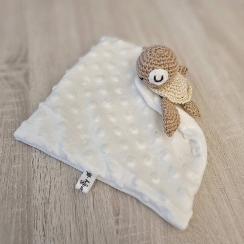May include: A white, textured baby blanket with a crocheted turtle toy. The turtle is brown and beige, with a smiling face. The blanket has a tag with the word "softly" on it.