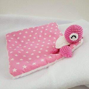 Peut inclure: Un jouet de tortue en crochet rose avec une carapace blanche posé sur une couverture à motifs d'étoiles roses et blanches.