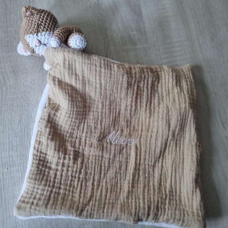 May include: A square-shaped baby security blanket made of tan crinkled muslin fabric with a white underside. The blanket features a crocheted brown and white cat head attached to one corner, with the name Meir embroidered in white script on the front.