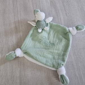 Peut inclure: Une couverture de sécurité pour bébé vert sauge avec une tortue en crochet attachée. La tortue a une tête et des ailes blanches et une carapace verte. Le nom "Naïm" est brodé en fil vert assorti.
