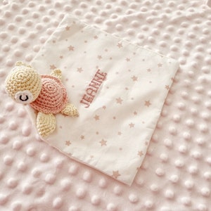 Peut inclure: Un jouet en crochet en forme de tortue rose et crème sur une couverture rose et blanche à motifs d'étoiles. La couverture est brodée du nom "Jennie".