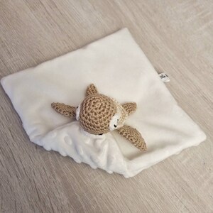 May include: A white, plush baby security blanket with a crocheted fawn head and arms. The fawn is light brown with white accents. The blanket has a textured, dotted pattern on the bottom half.