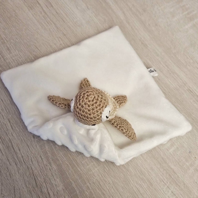 May include: A white, plush baby security blanket with a crocheted fawn head and arms. The fawn is light brown with white accents. The blanket has a textured, dotted pattern on the bottom half.