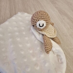 May include: A handmade crochet turtle toy in shades of tan and cream rests on a white, textured blanket. The turtle has a smiling face with black eyes and a white muzzle. The blanket has a soft, plush appearance.