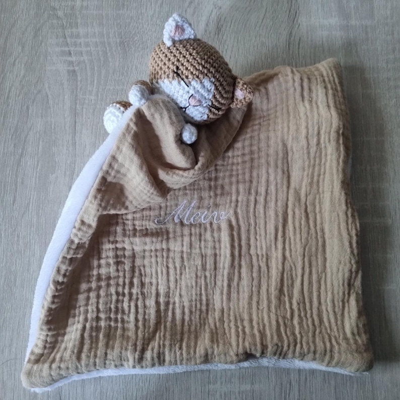 May include: A tan and white crochet cat security blanket featuring a soft, textured muslin fabric with the name "Meir" embroidered in white thread. The cat head has closed eyes and small ears, attached to a square blanket with a white underside.