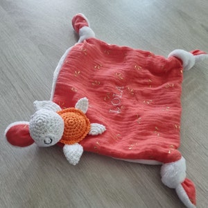 Peut inclure: Une couverture pour bébé de couleur corail avec une tortue en crochet blanc attachée. La couverture a une bordure blanche et des accents de paillettes dorées. Le nom "Lola" est brodé sur la couverture en blanc.