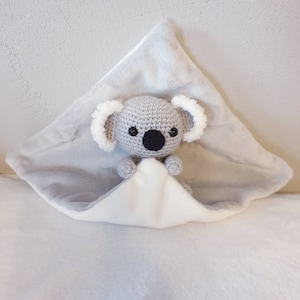 Puede incluir: Un koala de peluche gris tejido a crochet con ojos negros y una nariz negra está acurrucado dentro de una manta de felpa gris y blanca.
