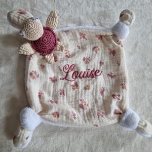 Peut inclure: Une couverture de sécurité pour bébé carrée en mousseline crème avec un motif floral rose. Le centre est brodé du prénom Louise en écriture rose. Une tortue crochetée avec une carapace rose foncé est fixée à l'un des coins.