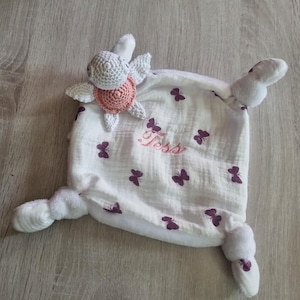 Peut inclure: Une couverture de sécurité pour bébé blanche avec une tortue crochetée et un motif papillon. La couverture a une texture douce et moelleuse et porte le nom "Tess" brodé en rose. La tortue a une tête, des ailes et une carapace roses.