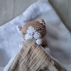 May include: A crocheted stuffed cat toy with light brown and white yarn, featuring closed eyes and a pink stitched nose. The cat is tucked under a textured beige muslin fabric blanket, with its small white paws visible resting on the fabric edge.