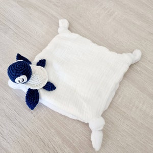 Peut inclure: Une couverture de sécurité blanche pour bébé avec une tête et des nageoires de tortue crochetées. La tortue a une tête et des nageoires bleu marine, avec une carapace blanche. La couverture est carrée avec des coins noués.
