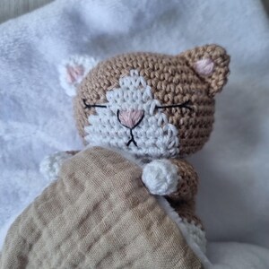 May include: A crocheted cat toy with light brown and white yarn, featuring closed eyes stitched in black thread, a pink nose, and pink inner ears. The cat is tucked under a textured, light brown fabric blanket with a visible grid-like pattern.