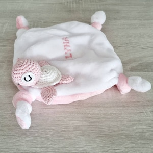 Peut inclure: Une couverture en peluche blanche et rose avec une tortue en crochet rose et blanche attachée. La couverture a une bordure rose et le texte "Emily" brodé dessus.