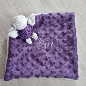 Peut inclure: Une couverture pour bébé violette avec une surface texturée à pois et une bordure blanche. Une tortue crochetée en blanc et violet est attachée à la couverture. Le nom "Alizee" est brodé en écriture blanche sur la couverture.