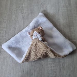 May include: A security blanket featuring a small, crocheted brown and white cat head with closed eyes and stitched features. The blanket has a soft white plush underside and a textured, light tan muslin fabric top layer folded over the cat.