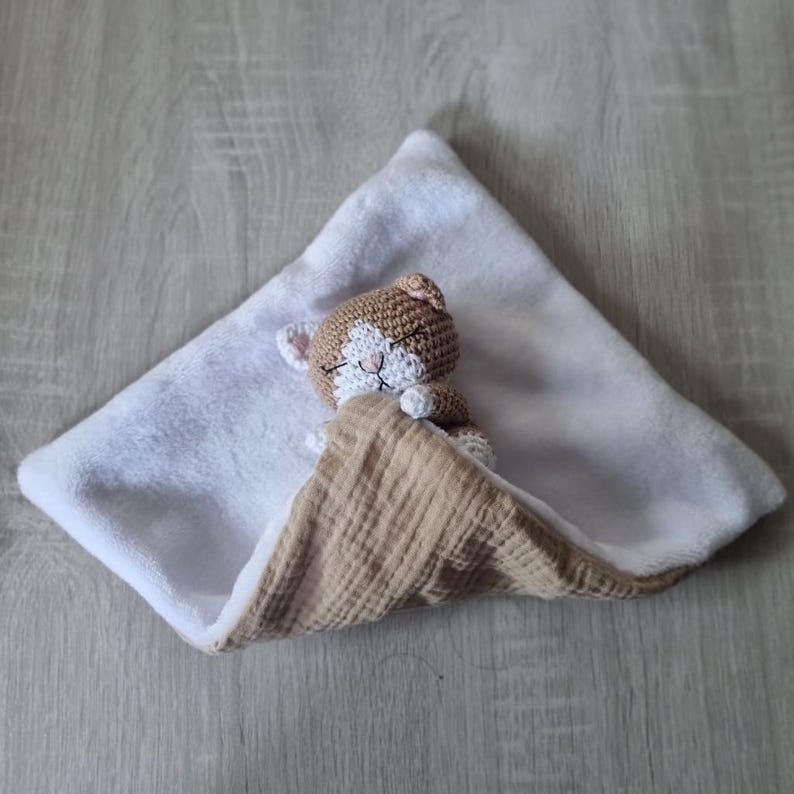 May include: A security blanket featuring a small, crocheted brown and white cat head with closed eyes and stitched features. The blanket has a soft white plush underside and a textured, light tan muslin fabric top layer folded over the cat.