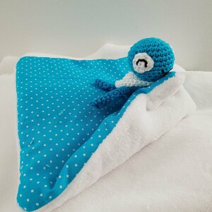 Peut inclure: Un animal en peluche bleu crocheté avec des yeux blancs et une bouche blanche, couché sur une couverture blanche à pois bleus.