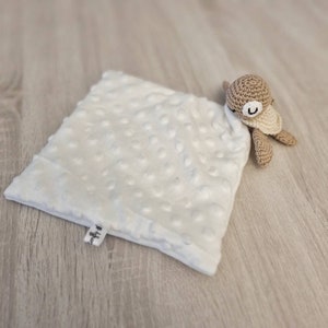 May include: A white, square baby lovey with a textured, dotted surface. A small, crocheted turtle in shades of tan and cream is attached to the corner. The lovey has a small tag with text.