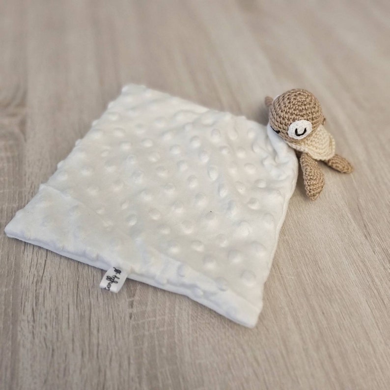 May include: A white, square baby lovey with a textured, dotted surface. A small, crocheted turtle in shades of tan and cream is attached to the corner. The lovey has a small tag with text.