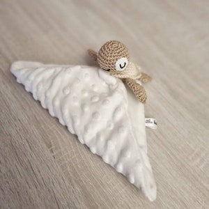 May include: A white, textured baby security blanket with a crocheted turtle toy attached. The turtle is tan and cream-colored with a smiling face. The blanket has a soft, plush feel and a small tag.