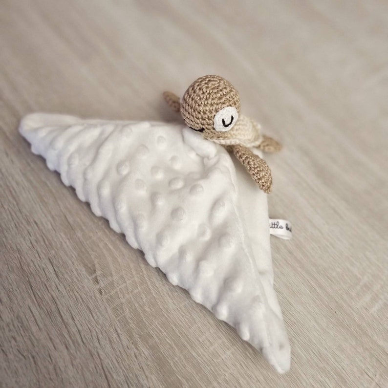 May include: A white, textured baby security blanket with a crocheted turtle toy attached. The turtle is tan and cream-colored with a smiling face. The blanket has a soft, plush feel and a small tag.