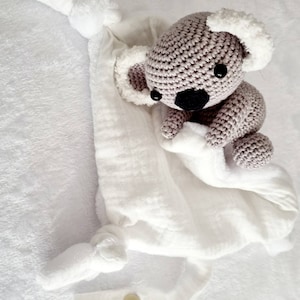 Puede incluir: Un koala de peluche gris tejido a crochet con orejas de piel blanca está unido a una manta de muselina blanca para bebé con un asa anudada y un cierre a presión.