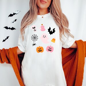 Peut inclure: Un t-shirt blanc avec une variété d'illustrations sur le thème d'Halloween, notamment un fantôme, une araignée, un crâne, une citrouille, une chauve-souris, une fiole de potion et du maïs sucré.