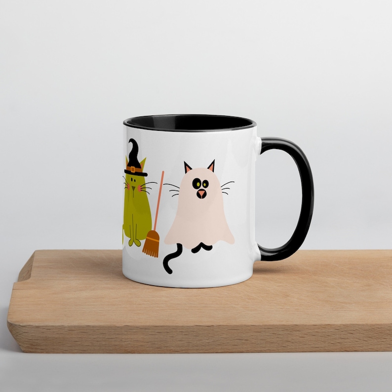 May include: White ceramic coffee mug with a black rim and handle. The mug features a green cat wearing a witch's hat and holding a broom, and a white ghost cat with black eyes.