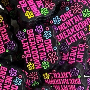 May include: Black, irregular-shaped accessories with the text "ONE MENTAL BREAKDOWN LATER" in pink. The accessories are decorated with colorful flower and dot designs in shades of yellow, green, blue, and purple.