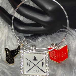 May include: A silver charm bracelet with a white square charm featuring the letters HP, a crossed wand, and a wizard's hat. The charm is surrounded by rhinestones. There are two additional charms: a gold wizard's hat and a red book with a lightning bolt and glasses.