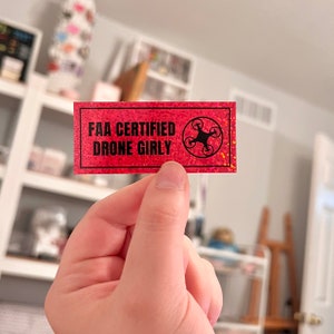 May include: A pink and glittery sticker with black text that reads "FAA CERTIFIED DRONE GIRLY" and a black silhouette of a drone with four propellers.