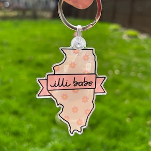 Può includere: Un portachiavi a forma dello stato dell'Illinois, rosa e bianco, con uno striscione rosa che recita "illi babe" e un motivo floreale.