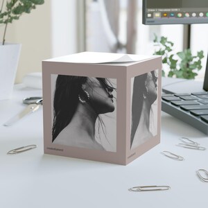 Puede incluir: Un bloc de notas de forma cúbica rosa con una foto en blanco y negro de una mujer con el pelo largo a cada lado. El texto "createdbykrist" está impreso en la parte inferior del bloc de notas.