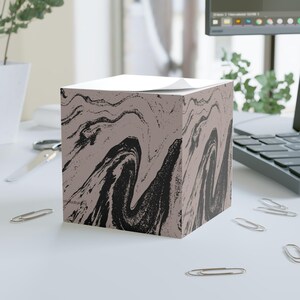 Puede incluir: Un portanotas cuadrado con un diseño de mármol negro y topo. El portanotas está sobre una superficie blanca con varios clips plateados. Un bloc de notas blanco está encima.