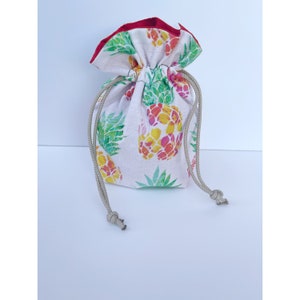 May include: A drawstring bag with a white background and a colorful pineapple pattern. The pineapples are yellow, pink, and orange with green leaves. The bag has a red trim and a beige drawstring.