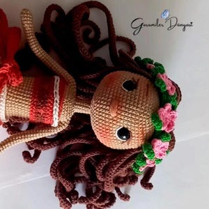 May include: A crocheted doll with brown hair, a red and white striped top, and a pink and green flower crown. The doll is lying on a white surface.