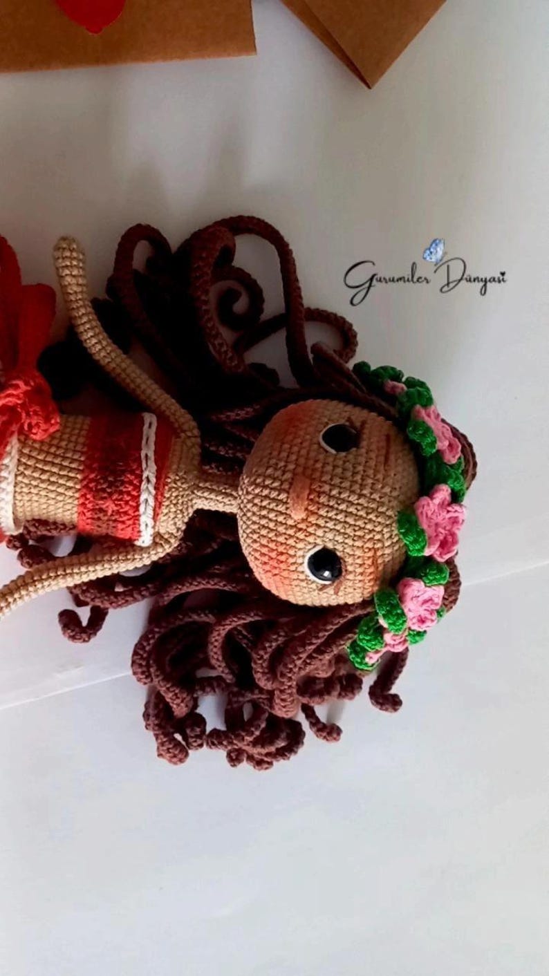 May include: A crocheted doll with brown hair, a red and white striped top, and a pink and green flower crown. The doll is lying on a white surface.