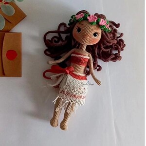 May include: A crocheted doll with brown hair, a flower crown, and a red and white dress. The doll is lying on a white surface.