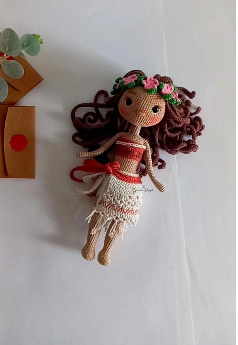 May include: A crocheted doll with brown hair, a flower crown, and a red and white dress. The doll is lying on a white surface.