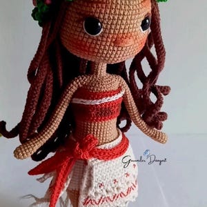 May include: Crocheted doll with brown hair, a flower crown, and a red and white dress. The doll is wearing a red sash and has a white crocheted skirt. The doll is standing on a white surface.