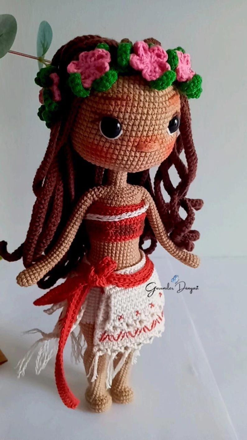 May include: Crocheted doll with brown hair, a flower crown, and a red and white dress. The doll is wearing a red sash and has a white crocheted skirt. The doll is standing on a white surface.