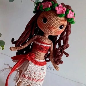 May include: Crocheted doll with brown hair, a red and white top, and a white skirt with red trim. The doll is wearing a flower crown and has a brown face with black eyes.