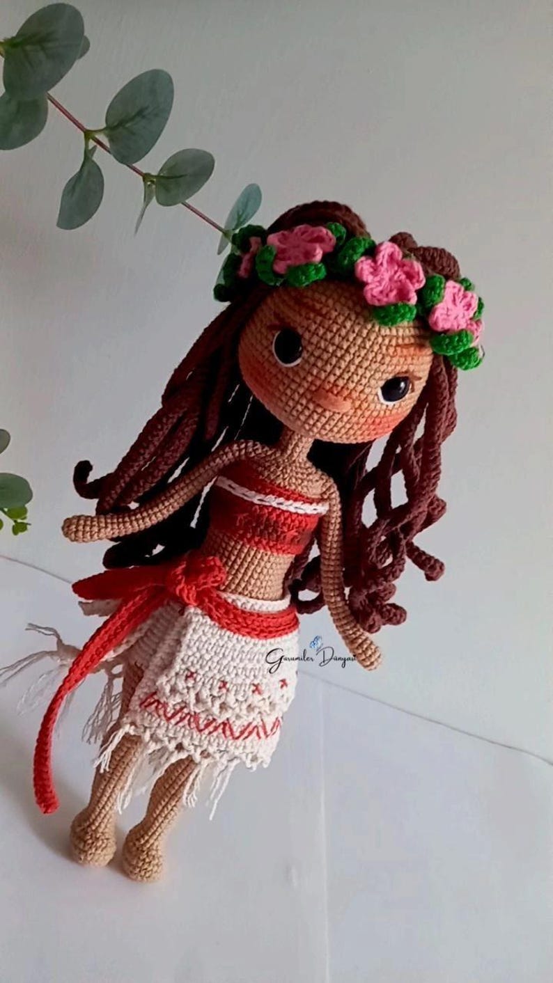 May include: Crocheted doll with brown hair, a red and white top, and a white skirt with red trim. The doll is wearing a flower crown and has a brown face with black eyes.