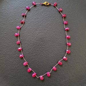 Op de afbeelding: Een delicate ketting met kleine, gefacetteerde, druppelvormige robijn edelstenen geregen op een dun, roze koord. De ketting heeft een goudkleurige sluiting. De robijnen zijn een levendige, dieproze kleur.