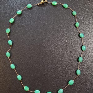May include: A delicate necklace featuring oval-shaped, light green gemstones strung on a thin, gold-colored chain. The necklace has a gold-colored clasp. The gemstones are evenly spaced along the chain, creating a simple yet elegant design.