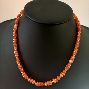 Peut inclure: Collier compos&eacute; de pierres pr&eacute;cieuses orange &agrave; facettes et de petites perles dor&eacute;es. Le collier est pr&eacute;sent&eacute; sur un buste de mannequin noir, mettant en valeur son design et sa couleur. Les pierres ont une teinte chaude et invitante.