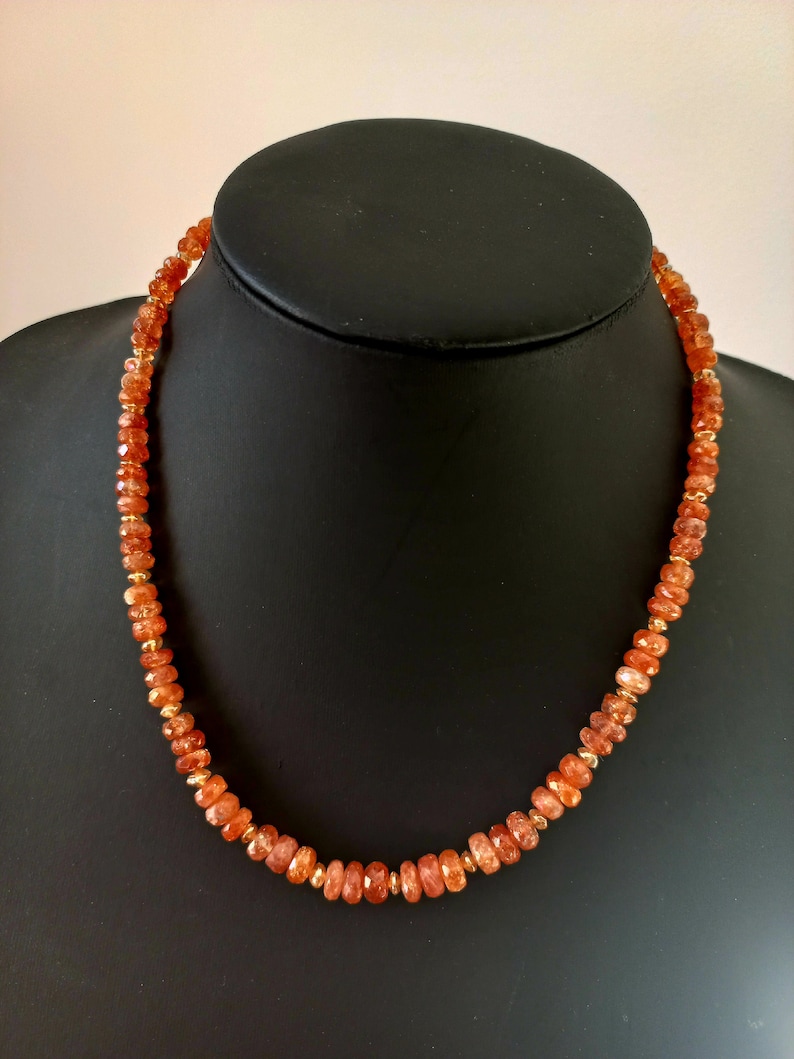 Peut inclure: Collier compos&eacute; de pierres pr&eacute;cieuses orange &agrave; facettes et de petites perles dor&eacute;es. Le collier est pr&eacute;sent&eacute; sur un buste de mannequin noir, mettant en valeur son design et sa couleur. Les pierres ont une teinte chaude et invitante.