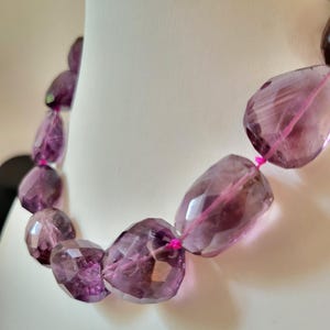 May include: A necklace featuring large, faceted, translucent purple amethyst beads strung together with a bright pink cord. The beads have an irregular, organic shape and a polished finish, creating a continuous chain of deep violet gemstones.