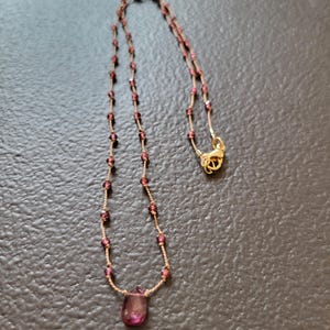 Puede incluir: Un collar delicado con un colgante de piedra preciosa en forma de lágrima. El collar está hecho de una fina cadena de oro con pequeñas cuentas de color rojo oscuro.